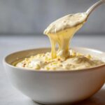 Close-up of a thick, rich cream sauce being lifted from a white bowl with a spoon, showing its smooth, stretchy texture.