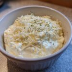 A white bowl filled with thick, rich cream sauce, generously topped with shredded cheese and dried herbs.