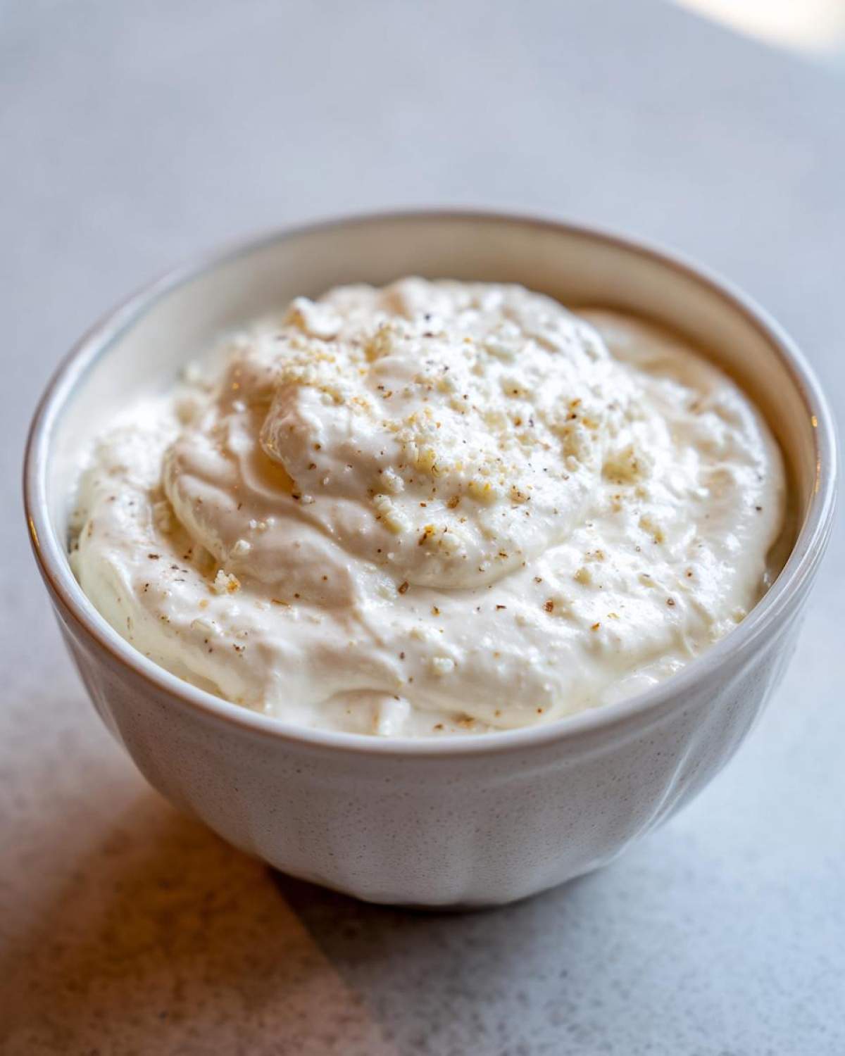 A close-up of a thick, rich cream sauce topped with grated cheese and pepper in a speckled white bowl.