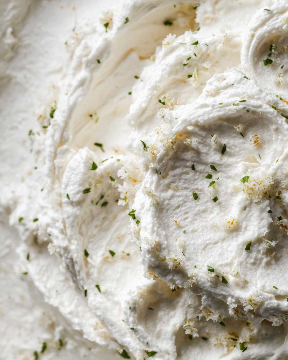 Macro shot showing the thick, swirled texture of a white cream sauce sprinkled with green herbs and zest.