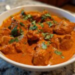 Close-up of rich, orange-red Crockpot Butter Chicken pieces garnished with fresh cilantro in a white bowl.