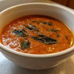 Close-up of creamy, orange-red tomato basil soup topped with dark green basil leaves in a white bowl.