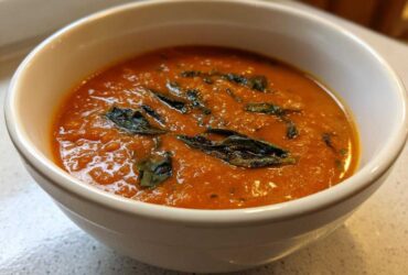 Close-up of creamy, orange-red tomato basil soup topped with dark green basil leaves in a white bowl.