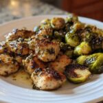 Close-up of a white plate featuring pieces of golden Sheet Pan Garlic Butter Chicken and roasted Brussels sprouts glistening with butter.