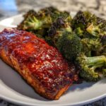 A close-up of a perfectly cooked salmon fillet topped with glossy honey harissa glaze next to roasted broccoli.