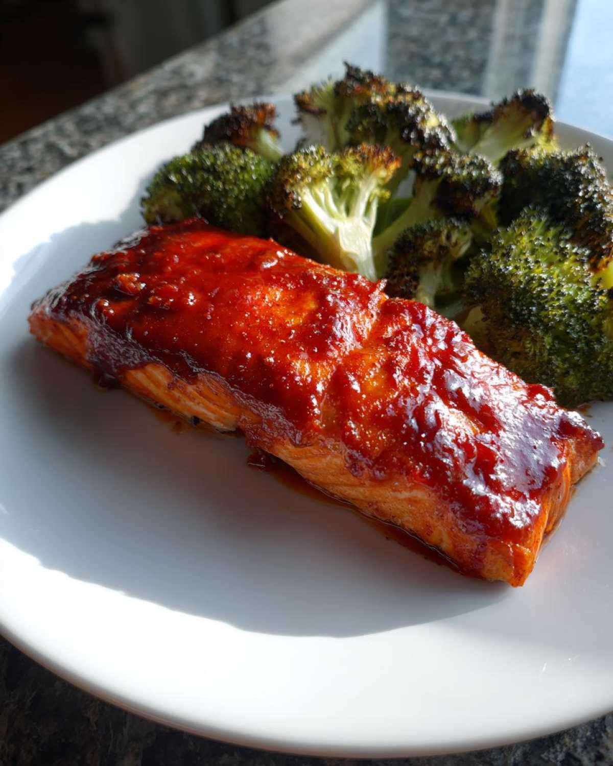 A serving of Sheet Pan Honey Harissa Salmon with a glossy red glaze next to roasted broccoli florets on a white plate.