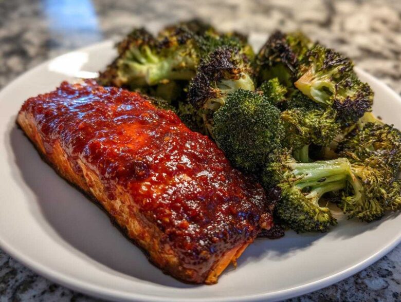 A close-up of a perfectly cooked salmon fillet topped with glossy honey harissa glaze next to roasted broccoli.