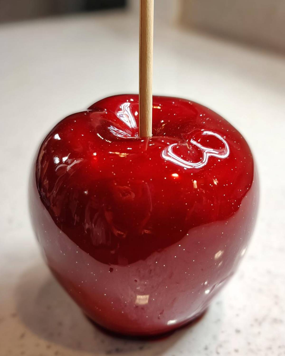 A close-up of a perfectly coated, glossy red candy apples with a wooden stick inserted.