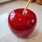 Close-up of a perfectly glossy, bright red candy apple with a wooden stick inserted, ready to eat.