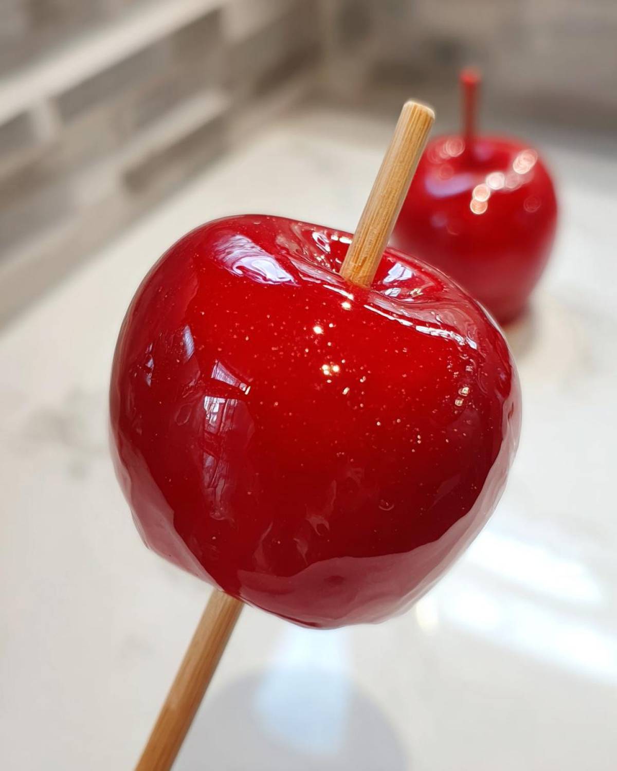 A close-up of a perfectly glazed, bright red candy apple on a wooden stick, with another candy apple blurred in the background.