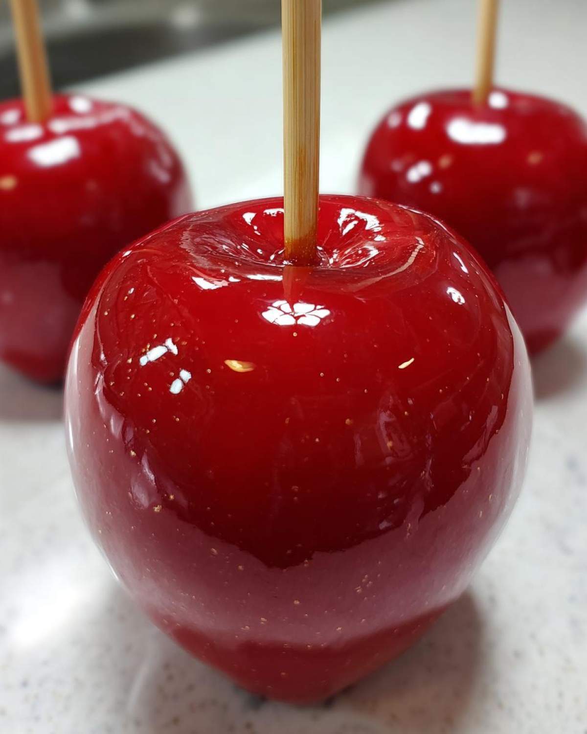 Close-up of a perfectly coated, shiny red candy apples on a wooden stick, with two more in the background.