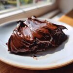 A close-up of glossy, rich chocolate ganache spread on a white plate, catching sunlight near a window.