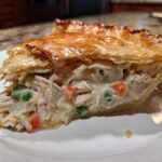 A generous slice of chicken pot pie recipe showing shredded chicken, peas, and carrots in a creamy sauce under a golden, flaky puff pastry crust.