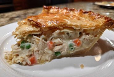 A generous slice of chicken pot pie recipe showing shredded chicken, peas, and carrots in a creamy sauce under a golden, flaky puff pastry crust.