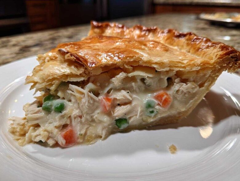 A generous slice of chicken pot pie recipe showing shredded chicken, peas, and carrots in a creamy sauce under a golden, flaky puff pastry crust.