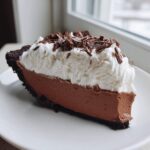 A decadent slice of french silk pie featuring a dark chocolate crust, rich chocolate filling, and topped with whipped cream and chocolate shavings.