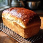 A golden brown, glossy loaf of soft bread cooling on a wire rack near a window.