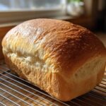 A golden brown, freshly baked loaf of soft bread cooling on a wire rack near a sunny window.