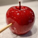 Close-up of a glossy, vibrant red candy apples with edible glitter and a wooden stick inserted.
