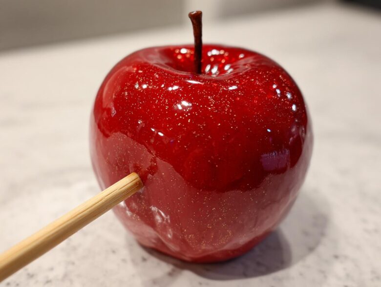 Close-up of a glossy, vibrant red candy apples with edible glitter and a wooden stick inserted.