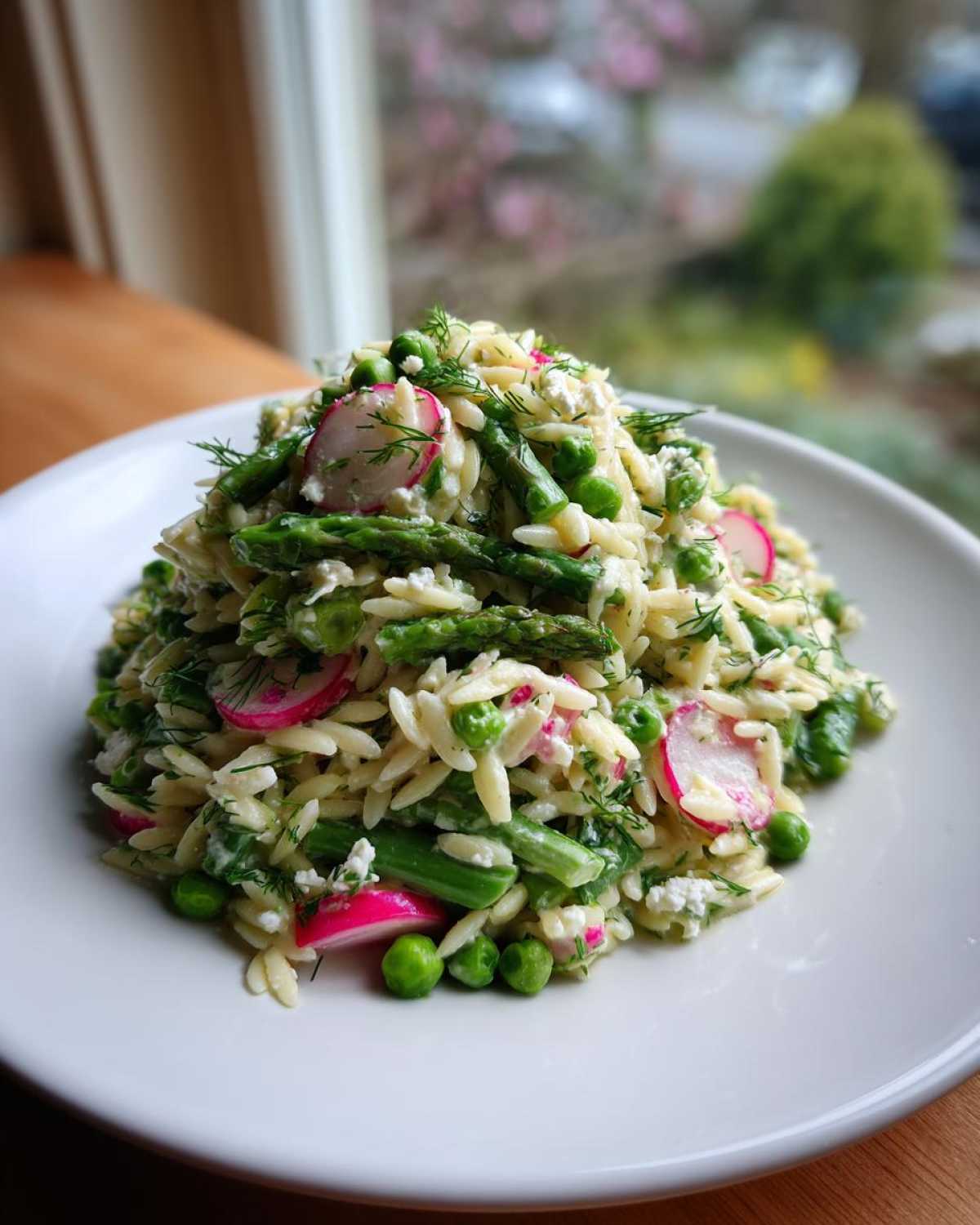 A mound of Spring Orzo Pasta Salad featuring bright green asparagus, peas, sliced radishes, and dill on a white plate.