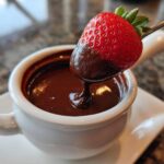 A fresh strawberry being dipped into a small white bowl of melted chocolate fondue.