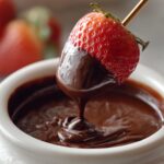 A fresh strawberry on a skewer being dipped into a white bowl of smooth, melted chocolate fondue.