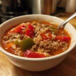 Close-up of a white bowl filled with steaming stuffed pepper soup featuring ground meat, rice, and chunks of red and green peppers.