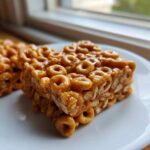 A close-up, tasty shot of a square of sticky, golden brown cheerio bars resting on a white plate.