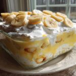 A glass dish filled with layers of THE BANANA PUDDING, topped with whipped cream and sliced bananas.