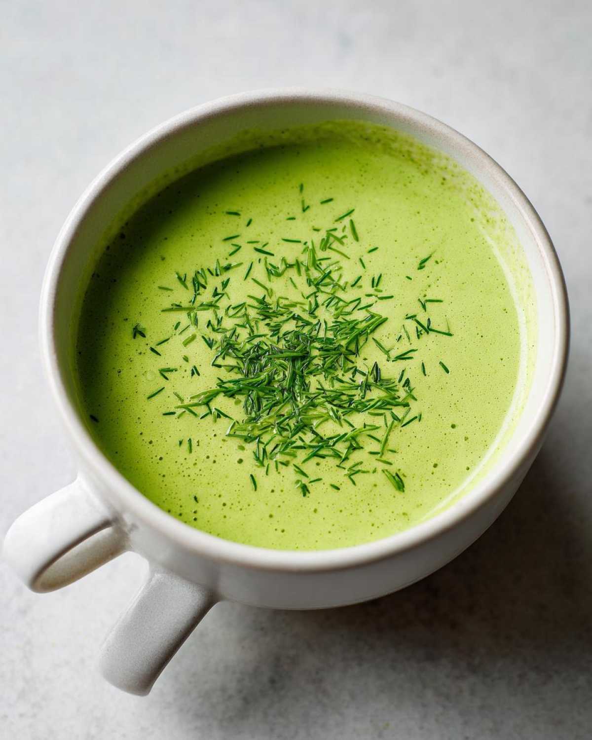 Overhead close-up of bright green Vegan Creamy Cucumber Dill Soup topped with fresh dill in a white mug.