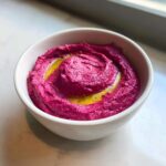 A close-up of vibrant magenta Vegan Creamy Roasted Beet Hummus swirled in a white bowl, topped with olive oil.