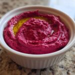 Close-up of vibrant pink Vegan Creamy Roasted Beet Hummus topped with olive oil in a white ramekin.