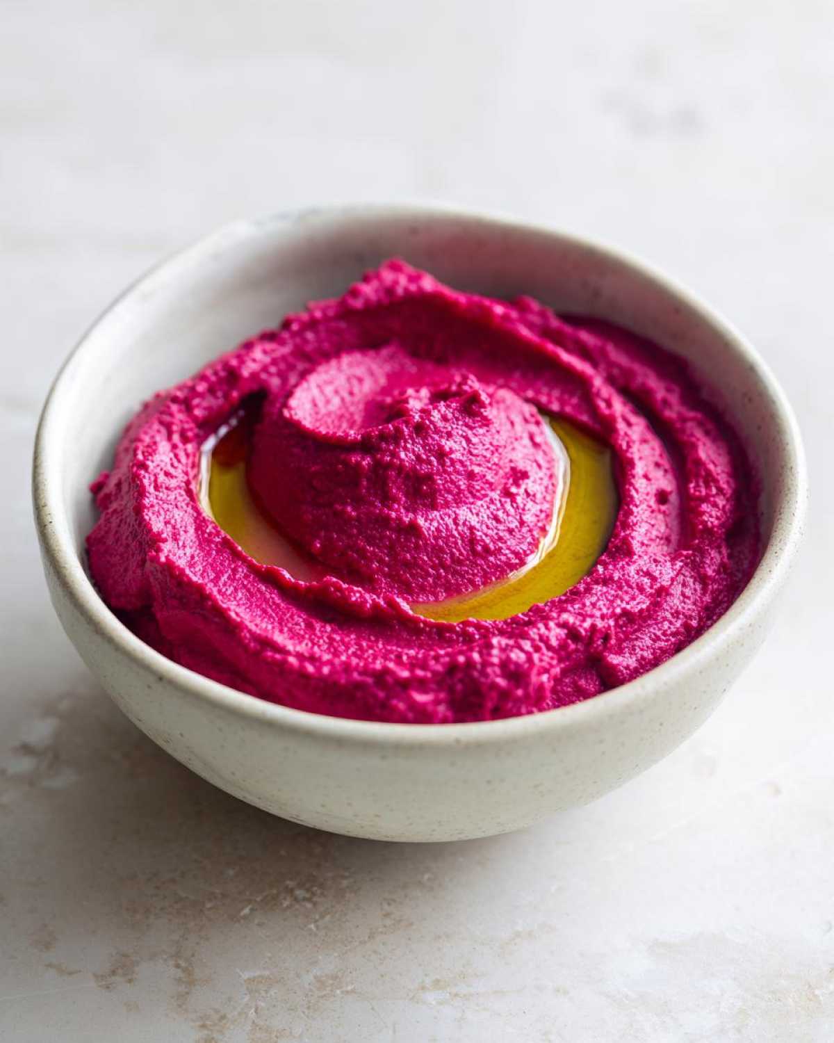Close-up of bright magenta Vegan Creamy Roasted Beet Hummus swirled in a light bowl, topped with olive oil.