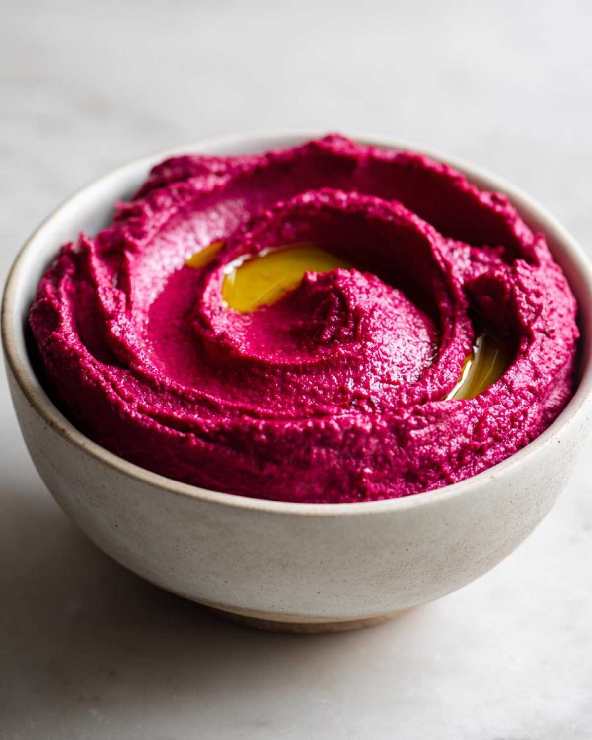Close-up of vibrant magenta Vegan Creamy Roasted Beet Hummus swirled in a light ceramic bowl, drizzled with olive oil.