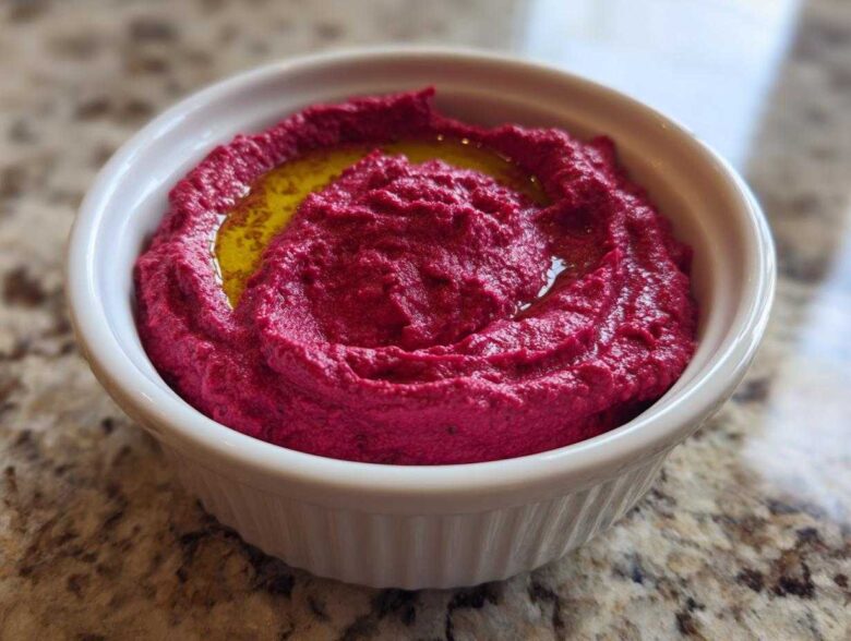 Close-up of vibrant pink Vegan Creamy Roasted Beet Hummus topped with olive oil in a white ramekin.