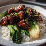 A close-up of a Vegan Crispy Sesame Tempeh Bowl featuring glazed tempeh pieces over white rice and wilted bok choy, sprinkled with sesame seeds.
