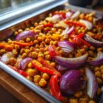 Close-up of seasoned chickpeas, red onion wedges, and red bell pepper strips roasting on a sheet pan for the Vegan One-Pan Chickpea Shawarma.