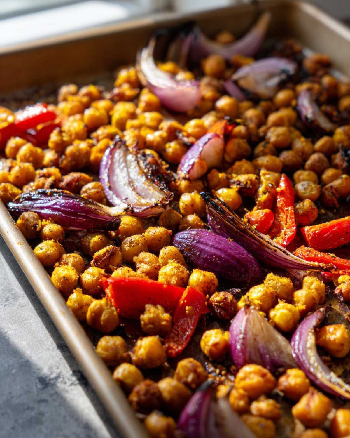 Close-up of roasted chickpeas, red onion wedges, and red pepper pieces on a sheet pan for Vegan One-Pan Chickpea Shawarma.