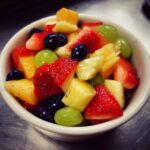 Close-up of a white bowl filled with colorful Fruit Salad featuring strawberries, blueberries, green grapes, and pineapple chunks.