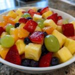 Close-up of a white bowl filled with colorful Fruit Salad, featuring strawberries, pineapple chunks, green grapes, and blueberries.