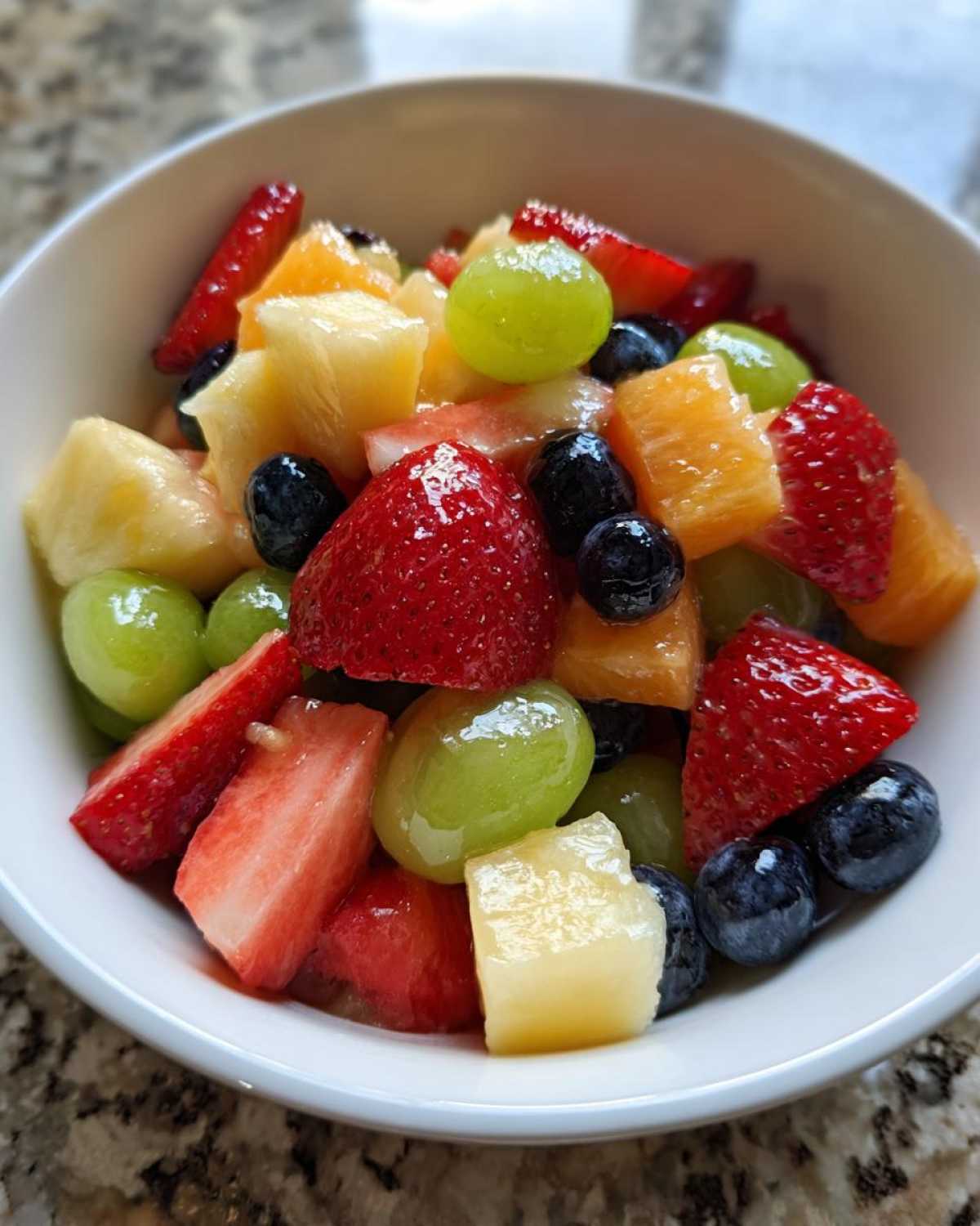 Close-up of a white bowl filled with a colorful Fruit Salad featuring strawberries, blueberries, pineapple chunks, and green grapes.