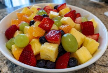 Close-up of a white bowl filled with colorful Fruit Salad, featuring strawberries, pineapple chunks, green grapes, and blueberries.