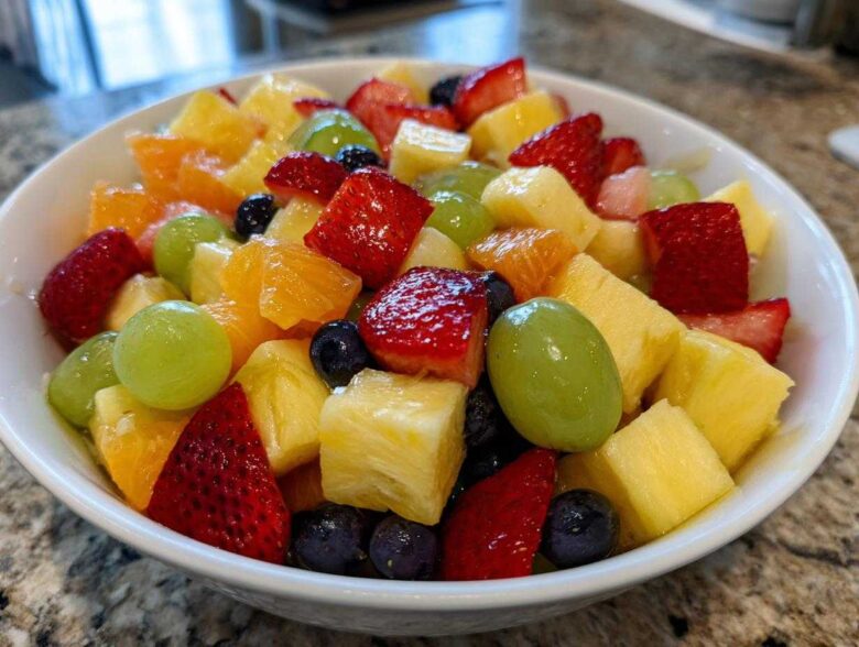 Close-up of a white bowl filled with colorful Fruit Salad, featuring strawberries, pineapple chunks, green grapes, and blueberries.