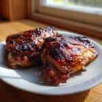 Two pieces of deeply glazed Baked Huli Huli Chicken thighs resting on a white plate, catching sunlight.