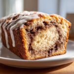 A close-up of a slice of Cinnamon Roll Banana Bread Loaf with a thick cinnamon swirl and white vanilla icing drizzled on top.