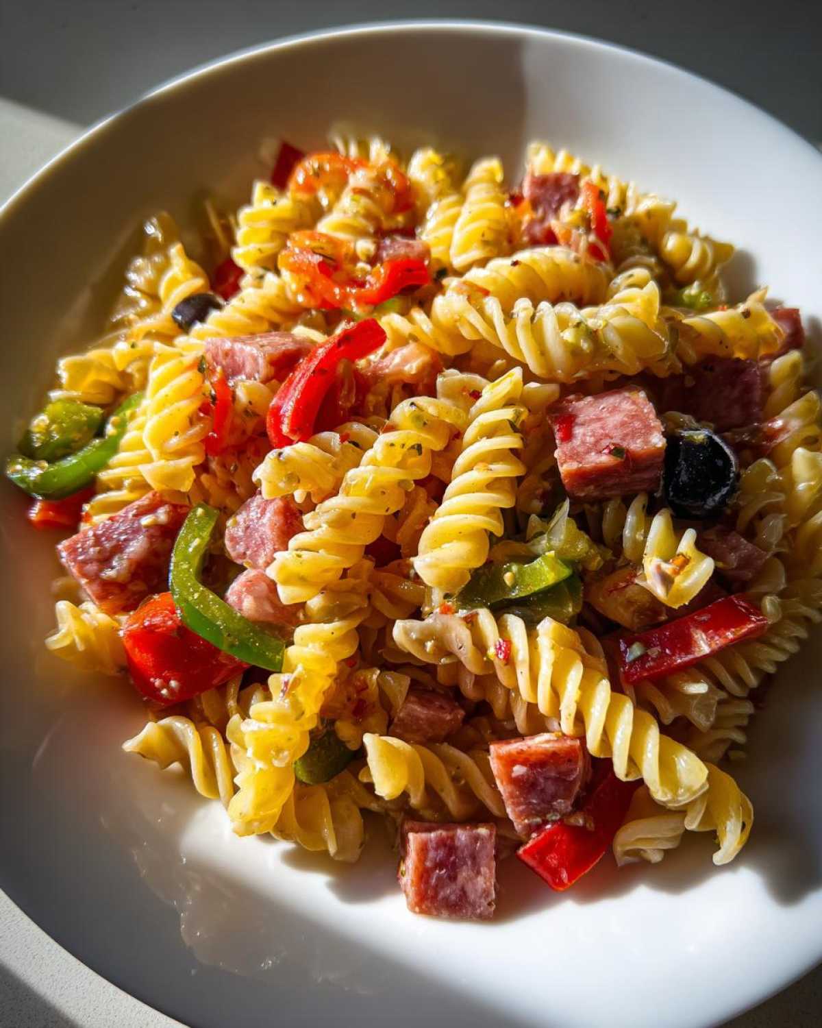 Close-up of Cold Pasta Salad with Italian Dressing featuring rotini pasta, cubed salami, red and green peppers, and black olives in a white bowl.