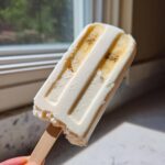 A creamy white Banana Coconut Popsicles featuring visible slices of banana embedded inside, held up near a window.