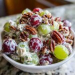 A close-up of a white bowl filled with vibrant red and green grapes coated in a creamy dressing, topped with toasted pecans, showcasing the delicious Grape Salad.