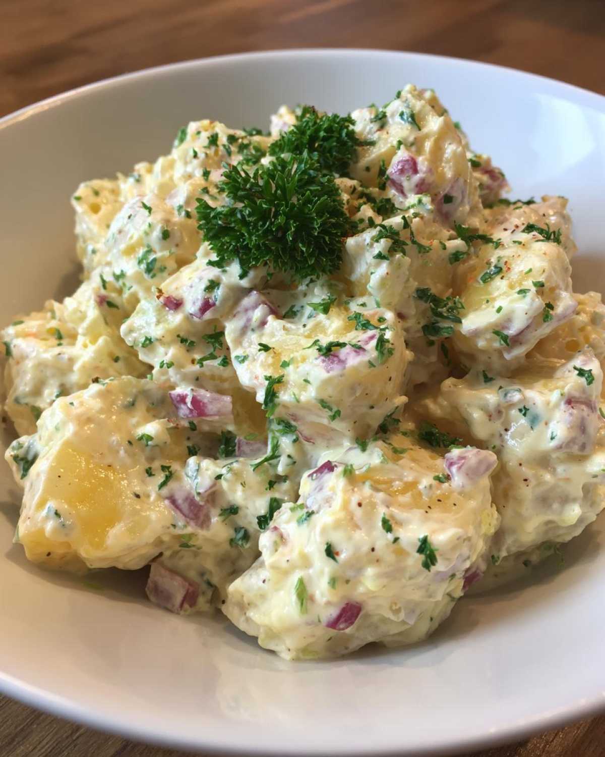 A close-up of a white bowl filled with rich, creamy potato salad featuring chunks of potato, red onion, and topped with fresh parsley.